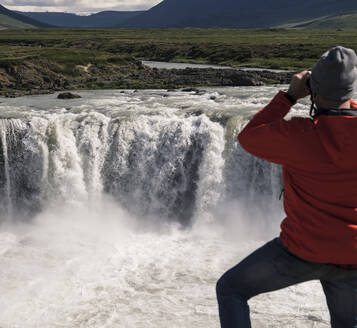 Man looking at Godafoss waterfalls, Iceland, with binoculars - UUF18795