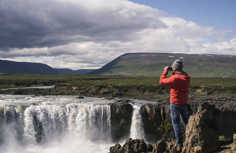 Mann betrachtet die Godafoss-Wasserfälle, Island, mit einem Fernglas, lizenzfreies Stockfoto