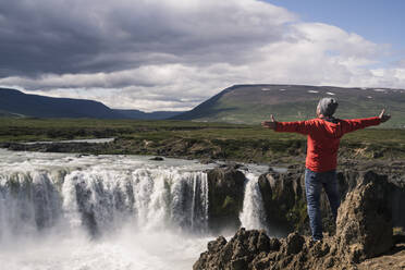 Mann mit Blick auf die Godafoss-Wasserfälle, Island, mit ausgestreckten Armen - UUF18797