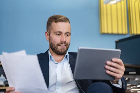 Geschäftsmann mit Papieren und Tablet am Schreibtisch im Büro - DIGF07956