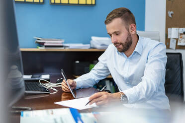 Geschäftsmann mit Papieren und Tablet am Schreibtisch im Büro - DIGF07959