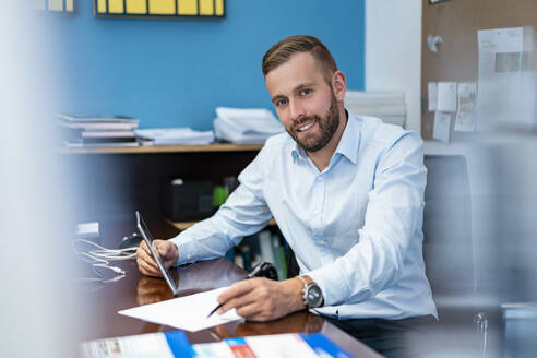 Portrait of smiling businessman with papers and tablet at desk in office - DIGF07960