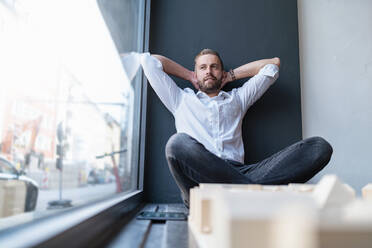 Entspannter Geschäftsmann mit Architekturmodell im Büro am Fenster sitzend - DIGF07975