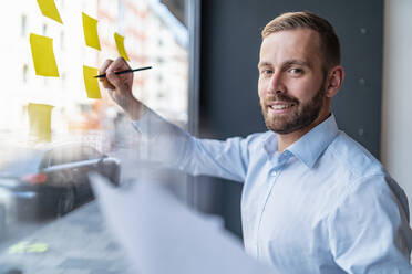 Portrait of smiling businessman writing on adhesive notes at glass pane - DIGF07990