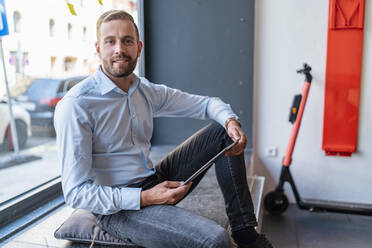 Portrait of smiling businessman with tablet at the window in office - DIGF07993