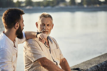 Vater und erwachsener Sohn sitzen auf einer Mauer am Flussufer und unterhalten sich - ZEDF02531