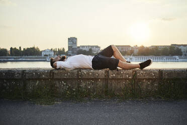 Man with beard lying on a wall at the riverside at sunset - ZEDF02538