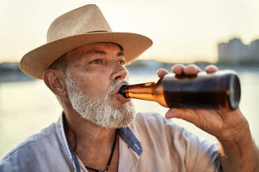 Senior man drinking a beer at the riverside at sunset - ZEDF02550