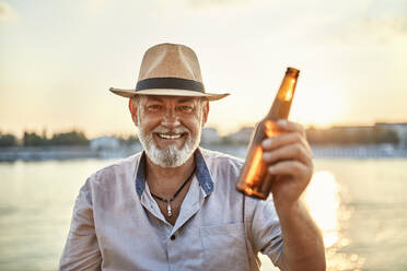 Porträt eines glücklichen älteren Mannes, der bei Sonnenuntergang am Flussufer ein Bier trinkt - ZEDF02551