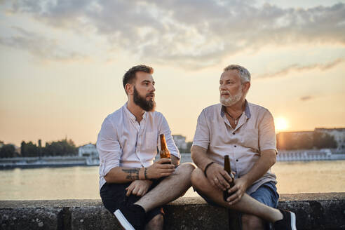 Father and adult son sitting on a wall at the riverside at sunset drinking a beer - ZEDF02554