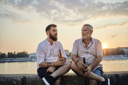 Vater und erwachsener Sohn sitzen bei Sonnenuntergang auf einer Mauer am Flussufer und trinken ein Bier - ZEDF02554