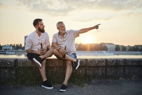 Vater und erwachsener Sohn sitzen bei Sonnenuntergang auf einer Mauer am Flussufer und trinken ein Bier, lizenzfreies Stockfoto