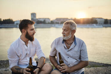 Vater und erwachsener Sohn sitzen bei Sonnenuntergang auf einer Mauer am Flussufer und trinken ein Bier - ZEDF02557