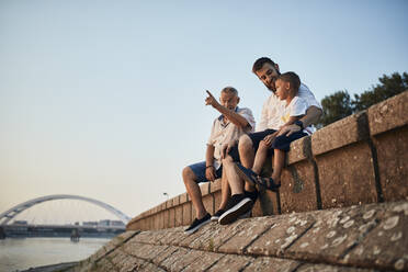 Großvater, Vater und Sohn sitzen auf einer Mauer am Flussufer - ZEDF02559