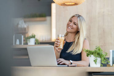 Porträt einer lächelnden blonden Geschäftsfrau mit Laptop und einem Glas Orangensaft in einem Café - DIGF08054
