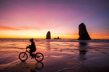 Silhouette eines kaukasischen Mädchens beim Radfahren in der Nähe von Felsformationen am Cannon Beach, Oregon, Vereinigte Staaten - BLEF14356