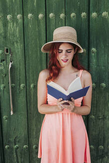 Portrait of redheaded young woman standing in front of green wooden door reading a book in summer - LJF00667