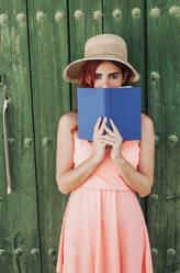 Portrait of redheaded young woman standing in front of green wooden door covering mouth with a book - LJF00668