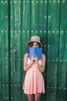 Portrait of redheaded young woman standing in front of green wooden door covering mouth with a book - LJF00669