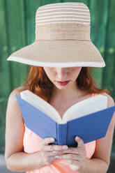 Rothaarige junge Frau vor grüner Holztür, die im Sommer ein Buch liest - LJF00672