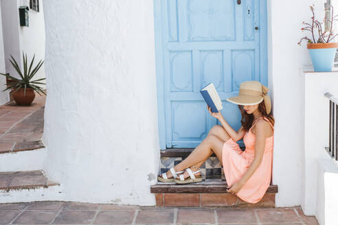 Junge Frau mit Buch auf der Treppe eines Hauseingangs sitzend, Frigiliana, Malaga, Spanien - LJF00691