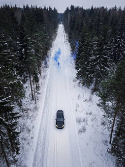 Luftaufnahme eines Mannes, der eine blaue Rauchgranate auf einer verschneiten Straße im Wald in Estland zündet. - AAEF02489