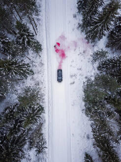 Aerial view of a man lighting a pink smoke grenade on a snowy road in the forest in Estonia. - AAEF02491