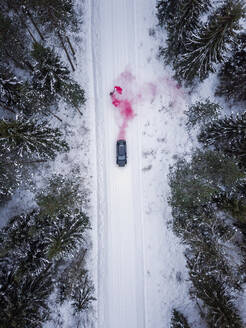 Luftaufnahme eines Mannes, der eine rosafarbene Rauchgranate auf einer verschneiten Straße im Wald in Estland zündet. - AAEF02491