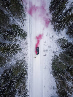 Luftaufnahme eines Mannes, der eine rosafarbene Rauchgranate auf einer verschneiten Straße im Wald in Estland zündet. - AAEF02493