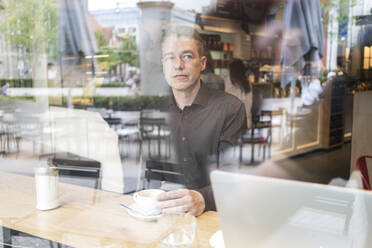 Mann sitzt in einem Café hinter einer Fensterscheibe, trinkt Kaffee mit Laptop - TAMF02050