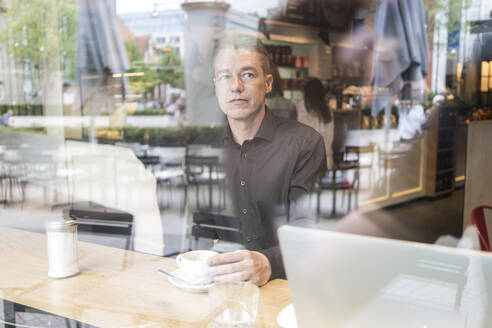 Mann sitzt in einem Café hinter einer Fensterscheibe, trinkt Kaffee mit Laptop - TAMF02050