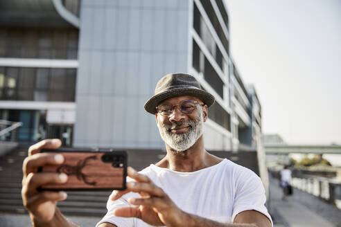 Porträt eines lächelnden reifen Mannes mit grauem Bart, der mit seinem Smartphone Selfies macht - FMKF05883
