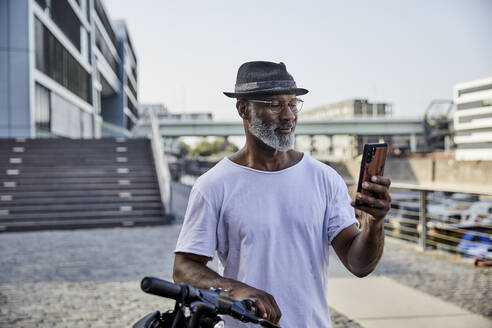 Porträt eines lächelnden reifen Mannes mit E-Scooter, der auf sein Mobiltelefon schaut - FMKF05886