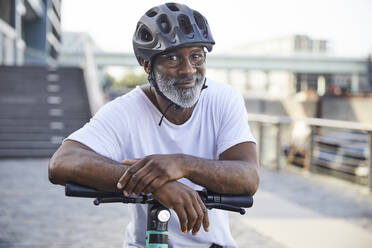Porträt eines lächelnden reifen Mannes mit Fahrradhelm, der sich auf den Lenker eines Elektrorollers stützt - FMKF05889