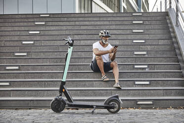 Älterer Mann mit E-Scooter, der auf einer Treppe sitzt und ein Mobiltelefon benutzt - FMKF05892