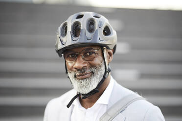 Portrait of smiling mature businessman with grey beard wearing cycling helmet and glasses - FMKF05896