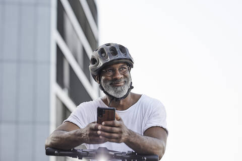 Porträt eines glücklichen reifen Mannes mit Fahrradhelm, der sich auf den Lenker eines Elektrorollers stützt, lizenzfreies Stockfoto