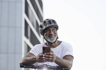 Porträt eines glücklichen reifen Mannes mit Fahrradhelm, der sich auf den Lenker eines Elektrorollers stützt - FMKF05902
