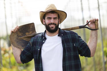 Portrait of smiling young man holding a shovel in a greenhouse - JSRF00509
