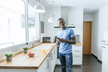 Porträt eines glücklichen jungen Mannes, der in der Küche zu Hause ein Mobiltelefon benutzt - KIJF02595