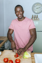 Porträt eines lächelnden jungen Mannes in der Küche, der eine Tomate für einen Salat schneidet - KIJF02599