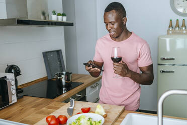 Junger Mann mit Weinglas und Mobiltelefon bei der Zubereitung von Salat - KIJF02606