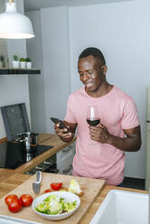 Smiling young man with wine glass using cell phone while preparing salad - KIJF02607