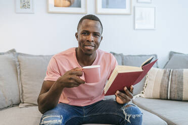 Porträt eines jungen Mannes mit Buch und Tasse Kaffee zu Hause - KIJF02609