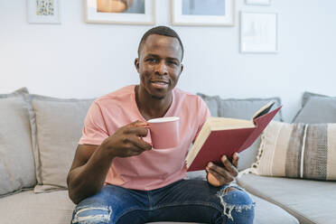 Portrait of young man with book and cup of coffee at home - KIJF02609