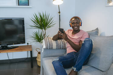 Portrait of smiling young man with headphones and smartphone on couch at home - KIJF02611