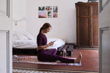 Young brunette woman during yoga, using smartphone in student dorm - IGGF01279