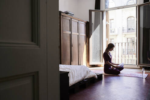Junge brünette Frau, die im Studentenwohnheim Yoga praktiziert und ein Buch liest - IGGF01288
