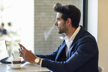 Businessman with smartphone and laptop in an urban cafe - JSMF01209