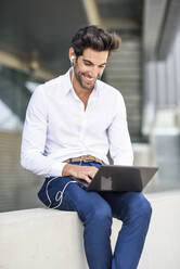 Lächelnder Geschäftsmann mit Kopfhörern, der einen Laptop im Freien in der Stadt benutzt - JSMF01237